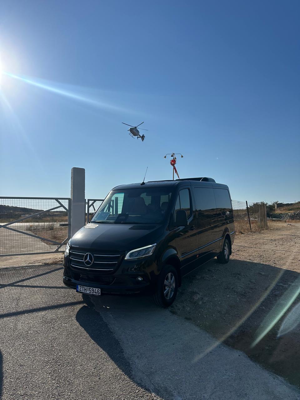 Black Mercedes-Benz Sprinter parked near a helipad gate with a helicopter airborne in the background under clear skies.