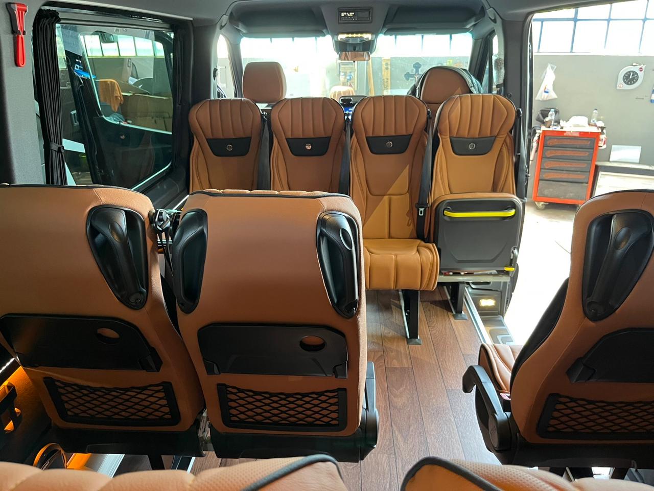 Interior cabin of the Travelbag VIP Mercedes Sprinter showing multiple rows of executive seating in warm tan leather with black piping, wood-effect flooring, foldable tray tables, and soft ambient lighting.