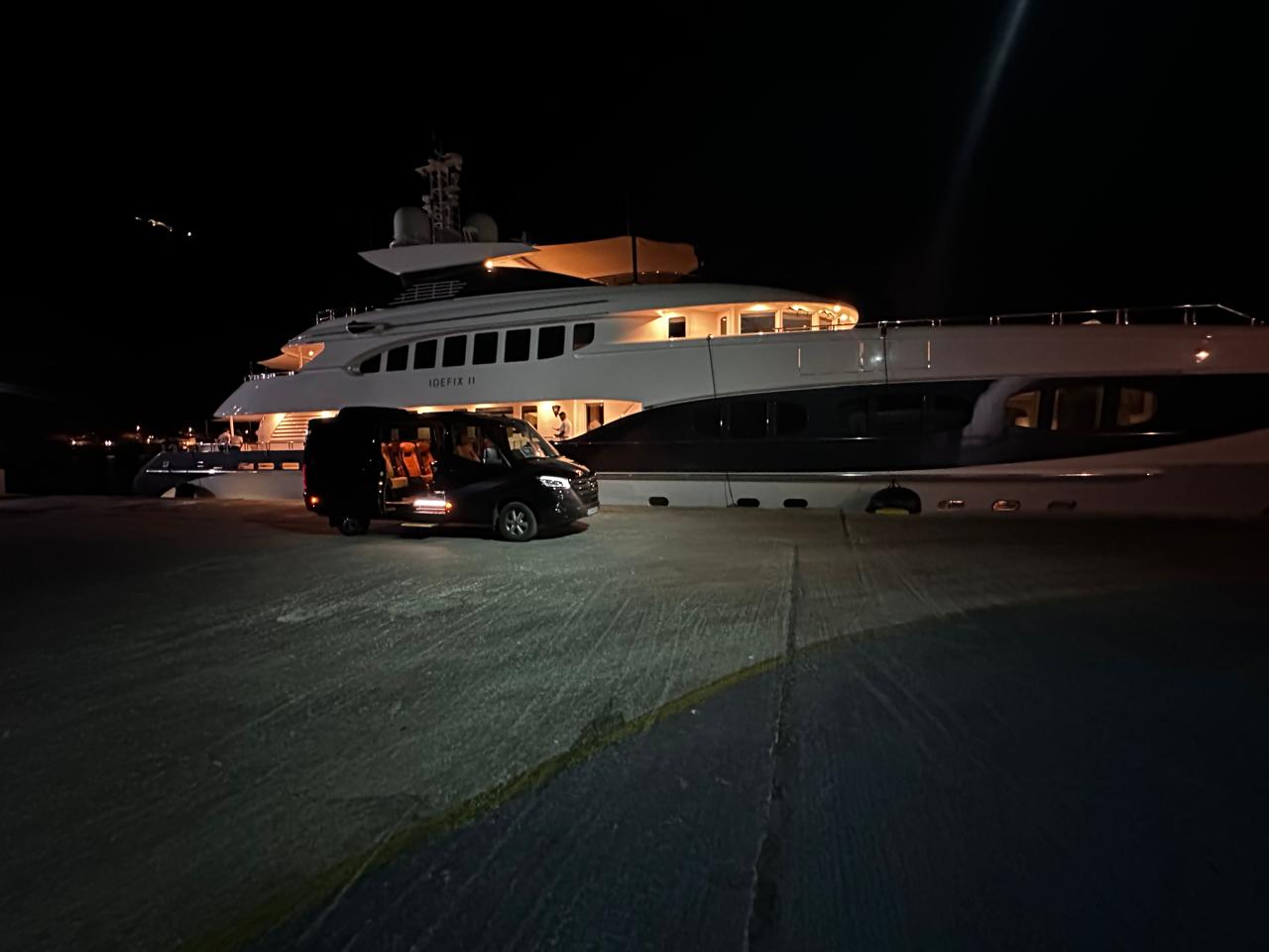 Black luxury VIP transfer van with illuminated interior parked alongside the superyacht IDEFIX II at a private marina dock at night