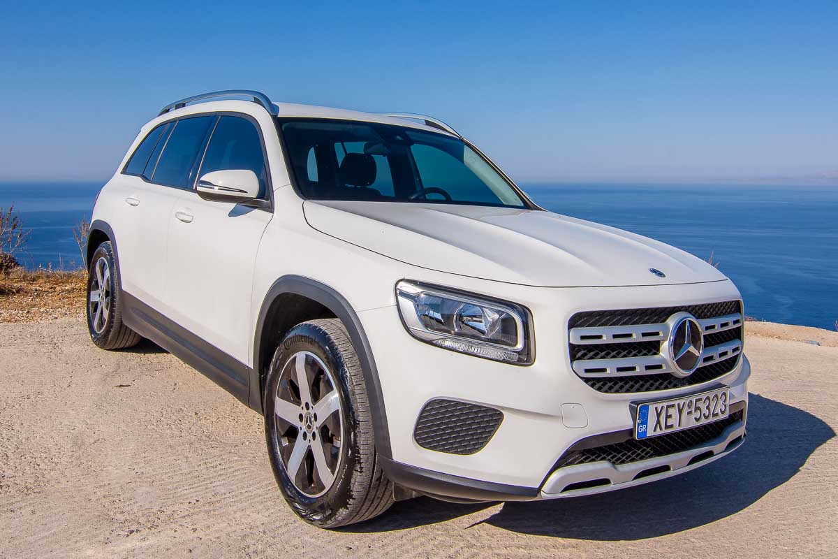 Front exterior view of a white Mercedes-Benz Sprinter luxury passenger van parked on a stone-paved terrace on a Sifnos hillside, overlooking the deep blue sea and a distant whitewashed Greek village.