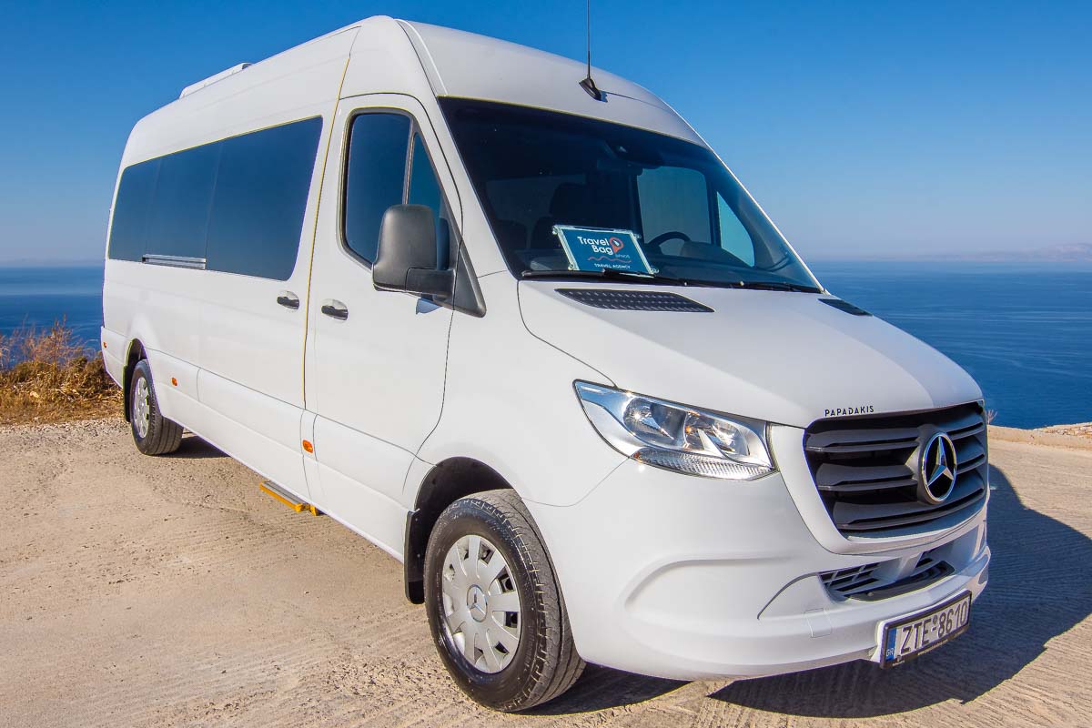 White Mercedes-Benz Sprinter passenger minibus parked at a sunny coastal overlook in Sifnos, with the deep blue sea and clear sky behind it.