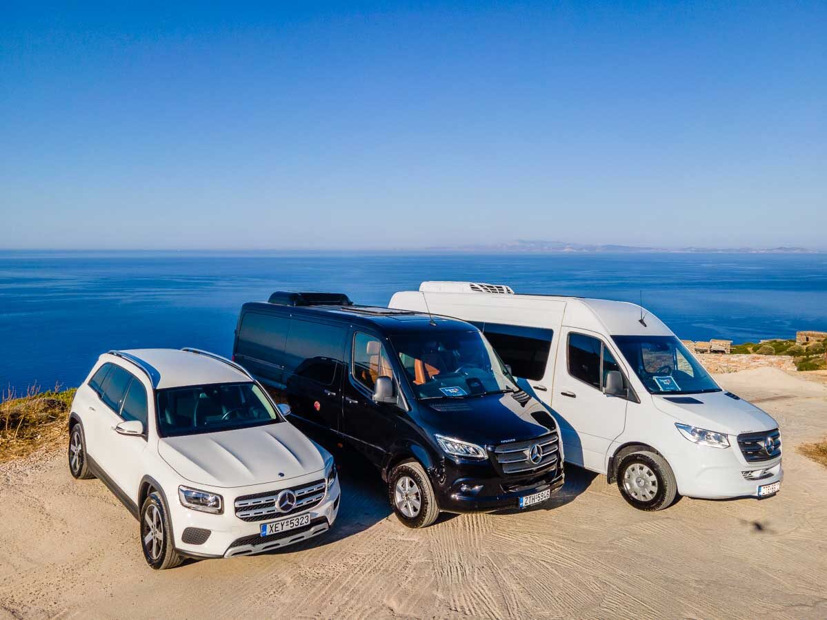 A white Mercedes-Benz SUV and a black Mercedes-Benz passenger van parked side by side near an open gate under a clear blue sky, part of the Travelbag Sifnos fleet.
